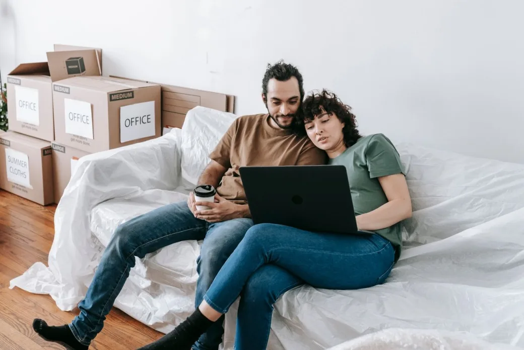Couple sitting on a covered couch among moving boxes, booking on a laptop