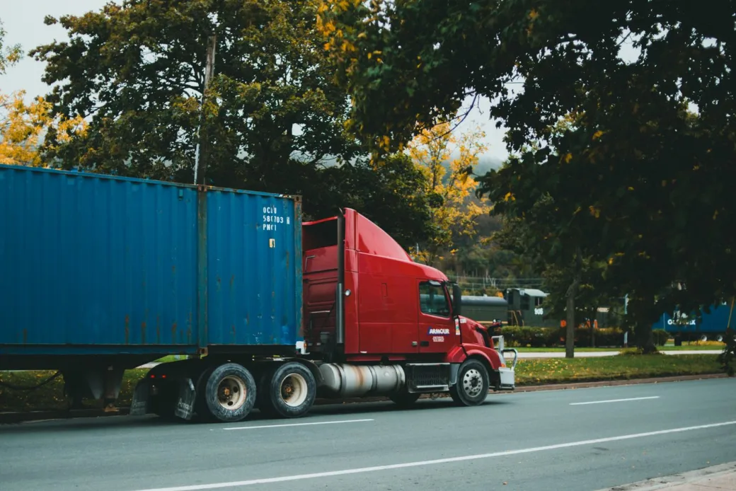 Red truck transporting a blue shipping container on a tree-lined road