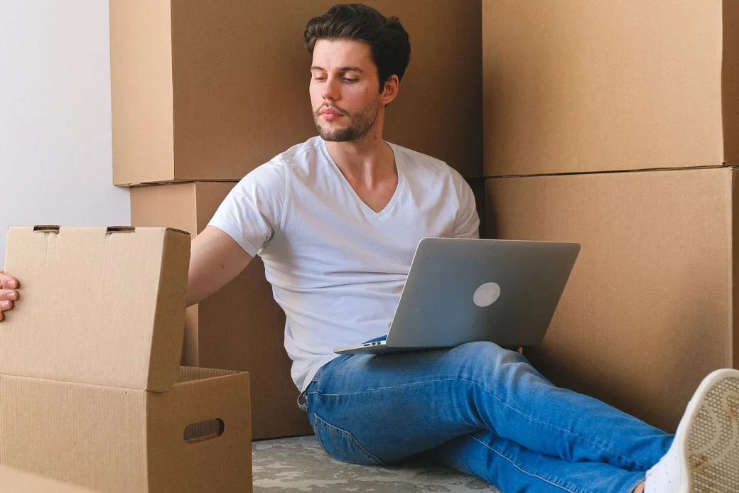 Person sitting on the floor at home with a laptop surrounded by moving boxes
